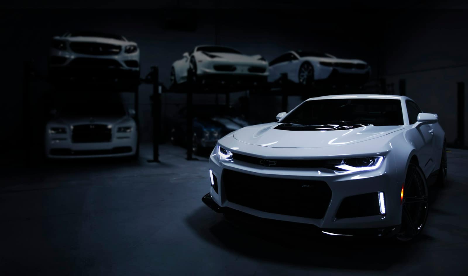 Loş galeri garajında liftlerde duran lüks araçlar, önde beyaz spor coupé